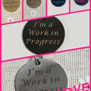 Pode incluir: Cinco etiquetas redondas de metal com o texto "I'm a Work in Progress" gravado nelas. As etiquetas são de diferentes cores: dourado, bronze, azul, arco-íris e prata.