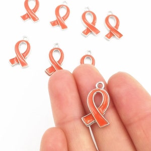 May include: Orange ribbon charms with silver-colored borders. The charms are in the shape of awareness ribbons, with a small loop at the top for attaching to jewelry or other crafts. The charms are on a white background.