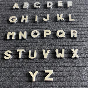 Puede incluir: Un conjunto de fichas de letras plateadas, dispuestas en orden alfab&eacute;tico, sobre un fondo de fieltro negro. Las fichas se utilizan para crear palabras y mensajes en un tablero de letras.