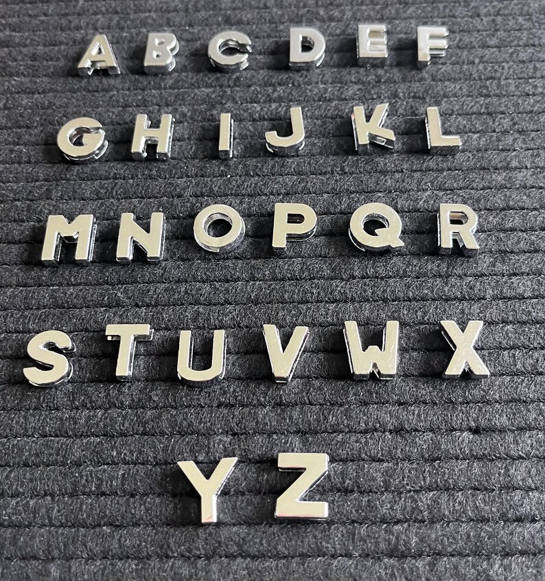 Puede incluir: Un conjunto de fichas de letras plateadas, dispuestas en orden alfab&eacute;tico, sobre un fondo de fieltro negro. Las fichas se utilizan para crear palabras y mensajes en un tablero de letras.