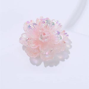 以下が含まれることがあります： 光沢のあるハイライトを備えた、薄いピンク色の半透明の花の形をした装飾品。花には複数の花びらの層があり、詳細な中心があります。このオブジェクトは、おそらく工芸品や装飾目的で使用されます。