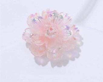 May include: A light pink, translucent flower-shaped embellishment with iridescent highlights. The flower has multiple layers of petals and a detailed center. The object is likely used for crafts or decorative purposes.