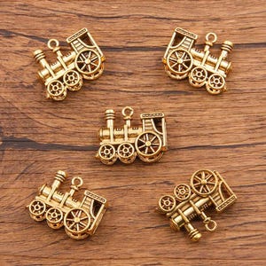 May include: Five gold-toned metal train engine charms. Each charm features a detailed design with wheels, a smokestack, and a cab. The charms are perfect for adding a whimsical touch to jewelry or crafts.