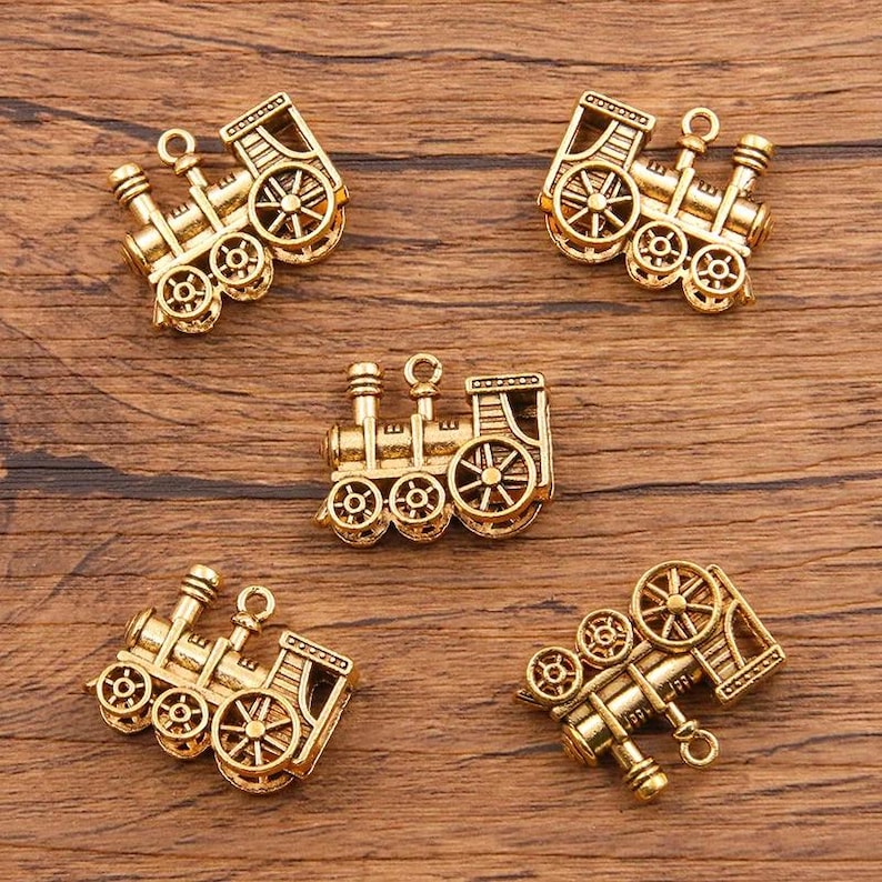 May include: Five gold-toned metal train engine charms. Each charm features a detailed design with wheels, a smokestack, and a cab. The charms are perfect for adding a whimsical touch to jewelry or crafts.