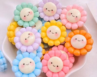 May include: A collection of colorful, smiling flower-shaped embellishments in various pastel shades, including blue, green, yellow, pink, and orange. Each flower has a white center with a cute face. These decorative items are likely for crafts or embellishments.