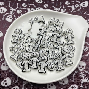 May include: A white plate filled with white and black mummy-shaped Halloween charms. The charms are made of plastic and have a glossy finish.