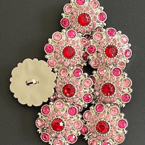 May include: A set of seven pink and red rhinestone buttons with silver settings. The buttons are arranged in a pyramid shape.