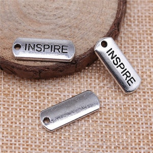 Könnte beinhalten: Drei silberfarbene Metall-Anhänger mit dem Wort "INSPIRE" auf zwei von ihnen graviert. Die Anhänger liegen auf einem gewebten Stoffhintergrund.
