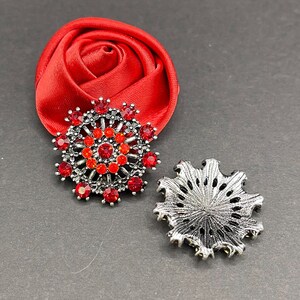 May include: Two silver brooches with red rhinestone accents. One brooch is a circular design with a center cluster of rhinestones. The other brooch is a starburst design with a center hole.