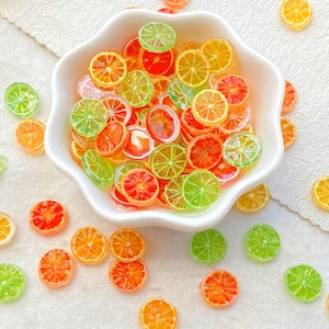 May include: A white, scalloped bowl filled with colorful, translucent fruit slices. The slices are orange, red, yellow, and green, resembling citrus fruits. Some slices are scattered around the bowl on a white surface.