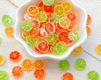 May include: A white, scalloped bowl filled with colorful, translucent fruit slices. The slices are orange, red, yellow, and green, resembling citrus fruits. Some slices are scattered around the bowl on a white surface.