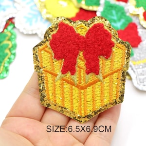 Puede incluir: Un parche de planchar cubierto de purpurina dorada con un diseño bordado en amarillo y rojo de una caja de regalo con un lazo rojo. El parche mide 6,5 x 6,9 cm.