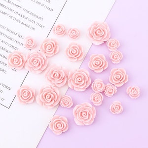 May include: A collection of 18 pink resin rose flower embellishments. The roses are in various sizes and are scattered on a light purple and white surface.