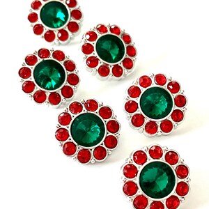 May include: Six decorative buttons with a silver base, featuring a large, round, emerald-green center stone surrounded by smaller, red, round stones. The buttons are arranged on a white background.