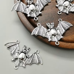 May include: A set of silver-toned metal bat charms, each featuring a detailed wing structure and a small circular loop at the top and bottom for attachment. The charms have a polished finish with dark recessed areas to highlight the bat shape.