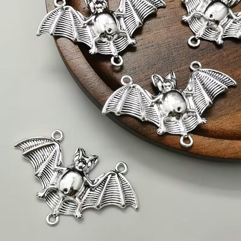 May include: A set of silver-toned metal bat charms, each featuring a detailed wing structure and a small circular loop at the top and bottom for attachment. The charms have a polished finish with dark recessed areas to highlight the bat shape.