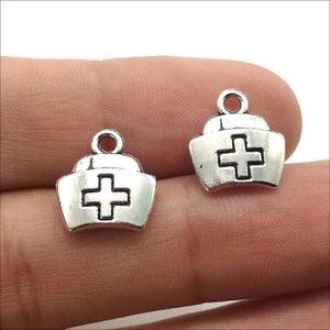 May include: Two silver-toned nurse hat charms with a white cross in the center. The charms have a loop at the top for attaching to a necklace or bracelet.