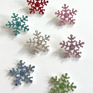 May include: A collection of sparkling snowflake brooches in various colors, including red, teal, pink, silver, blue, and green. Each brooch is adorned with multiple crystals, creating a dazzling effect. These decorative pins are perfect for winter-themed accessories.