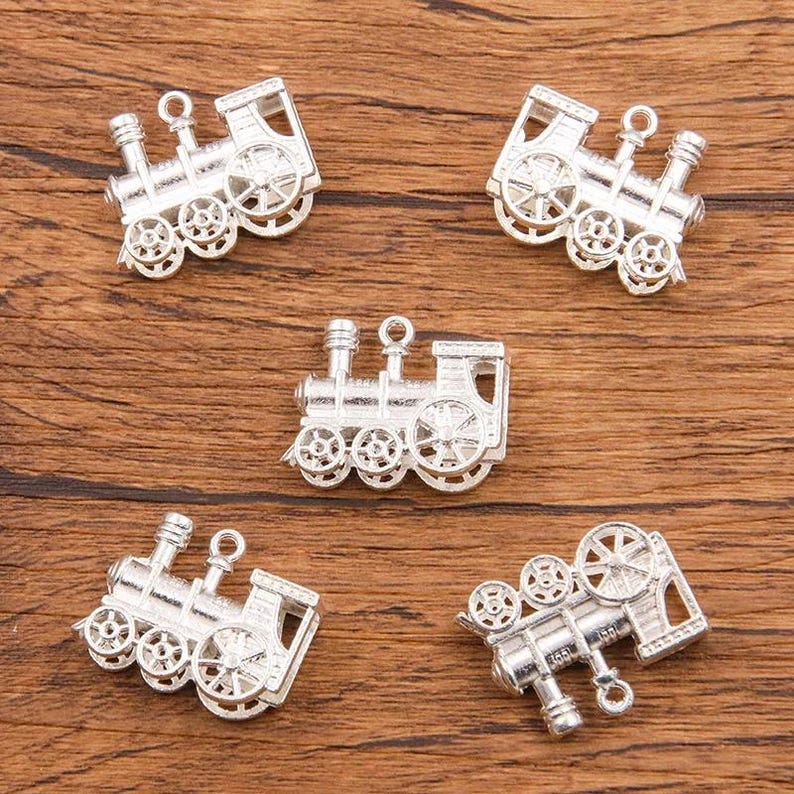 May include: Five silver-toned metal train charms with intricate details. Each charm features a locomotive with wheels, a smokestack, and a bell.