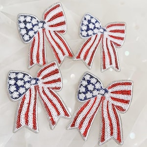 May include: Four decorative bow-shaped patches featuring a sequined American flag pattern. Each bow displays a blue section with white star-like shapes and alternating red and white stripes, all outlined with a silver border on a fabric base.