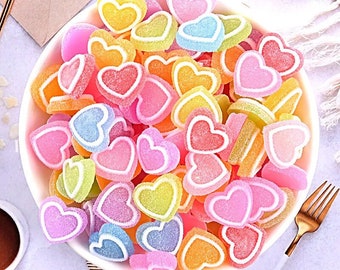 May include: A white bowl filled with colorful heart-shaped gummies. The gummies are in shades of pink, blue, yellow, and orange.