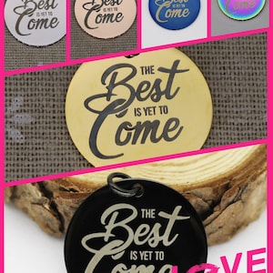 May include: A set of five round metal charms with the phrase "The Best Is Yet To Come" engraved in a cursive font. The charms are in different colors: silver, rose gold, blue, rainbow, and gold. The charms are displayed on a wooden surface with a pink background that says "Love Story".