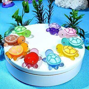 Puede incluir: Una colección de coloridas figuras de tortugas de cristal en varios colores, incluyendo verde, morado, rosa, azul, amarillo, naranja y rojo. Las tortugas están dispuestas sobre una plataforma circular blanca, con un fondo azul y acentos de plantas verdes.