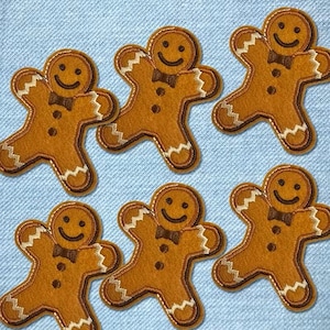 以下が含まれることがあります： 笑顔、蝶ネクタイ、ボタンのディテールが特徴の、6つのジンジャーブレッドマンパッチ。パッチは茶色で、タンと白のトリムが付いており、水色の生地の背景に配置されています。ホリデー工芸に最適です。