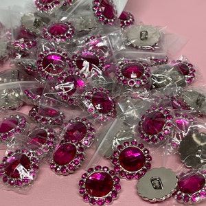May include: A large group of pink rhinestone buttons with silver settings. The buttons are arranged in a random pattern on a pink surface.