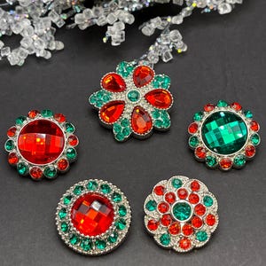 May include: Five decorative Christmas ornaments with red and green rhinestones. The ornaments are round and have a silver metal base.