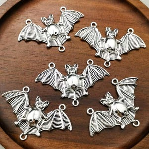 May include: Five silver-toned metal bat charms, each featuring a detailed body and spread wings. Every charm includes two small circular loops at the top and bottom, designed for connecting to jewellery components or other craft projects.