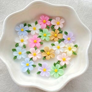 May include: A collection of colorful, miniature flower embellishments in a white, scalloped dish. The flowers are in shades of pink, yellow, blue, green, and white, each with a yellow center and green leaves. These decorative flowers are ideal for crafts.
