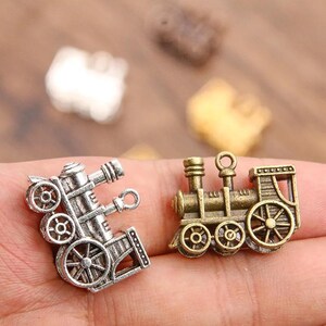 May include: Two silver and bronze train engine charms. The silver charm has a detailed design with wheels and a smokestack. The bronze charm has a more simplified design with wheels and a smokestack.