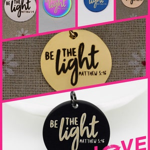 Puede incluir: Un conjunto de cinco dijes redondos de metal con la frase "Sé la luz Mateo 5:16" en letras negras. Los dijes son de diferentes colores: blanco, iridiscente, azul, dorado y negro.