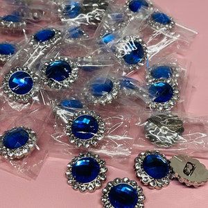 May include: A close-up of a pile of blue and silver rhinestone buttons. The buttons are round and have a single large blue rhinestone in the center surrounded by smaller silver rhinestones.