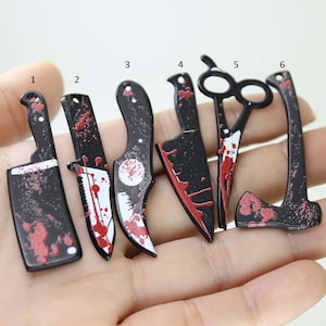 May include: Six black metal charms shaped like knives, an axe, and a pair of scissors. The charms are decorated with red paint to look like blood splatters.