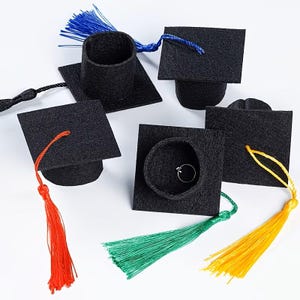 Può includere: Una collezione di piccoli cappelli di laurea neri con nappe colorate. I cappelli sono quadrati con una base cilindrica e le nappe sono in vari colori, tra cui rosso, verde, giallo e blu. Un cappello contiene un anello d'argento.