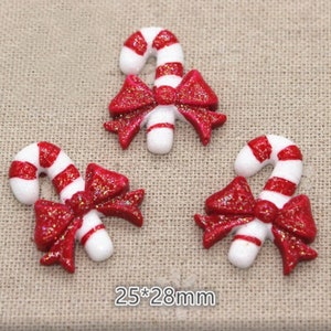 May include: Three Christmas-themed candy cane ornaments. Each ornament features a white candy cane with red stripes and a red glittery bow. The ornaments measure 25mm by 28mm.