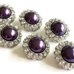 May include: Six decorative buttons with a central, round, dark purple faux pearl surrounded by a halo of clear, sparkling rhinestones. The buttons have a silver-colored metal base and are arranged on a white surface.