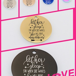 May include: A set of six round metal charms with the phrase "let her sleep for when she wakes she will move mountains" engraved on them. The charms are in different colors, including silver, gold, blue, and rainbow.
