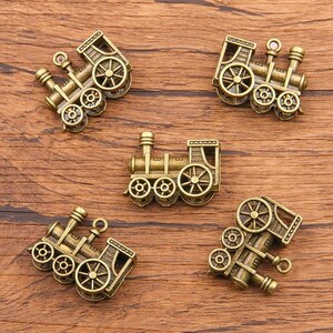 May include: Five antique gold-toned train engine charms. Each charm features a detailed design with wheels, a smokestack, and a bell. The charms are perfect for adding a vintage touch to jewelry or crafts.