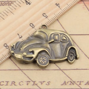 May include: A bronze-colored metal charm shaped like a vintage car. The charm has a detailed design with a rounded hood, a windshield, and two doors. The car has four wheels and a small loop at the top for attaching to a necklace or bracelet.