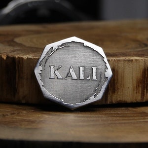 May include: A silver-colored, octagonal-shaped metal token with the word "KALI" engraved in the center. The token has a brushed metal finish and a slightly distressed edge.