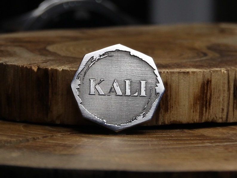 May include: A silver-colored, octagonal-shaped metal token with the word "KALI" engraved in the center. The token has a brushed metal finish and a slightly distressed edge.