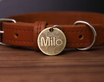May include: A gold-colored dog tag with a circular pattern and the name "Milo" engraved on it. The tag is attached to a brown leather dog collar.