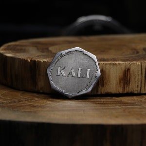 May include: A silver-colored, octagonal-shaped metal token with the word "KALI" engraved in the center.
