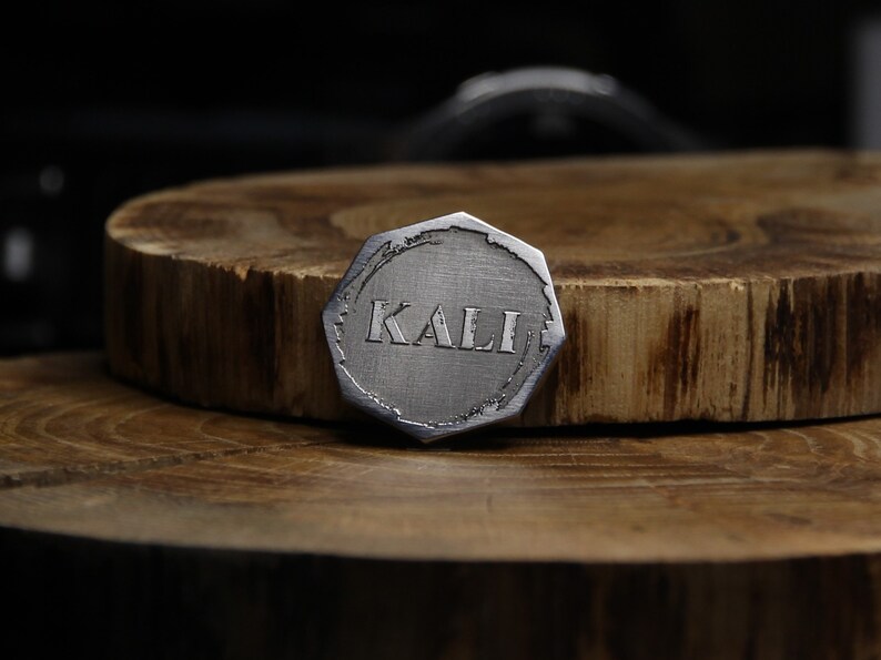 May include: A silver-colored, octagonal-shaped metal token with the word "KALI" engraved in the center.