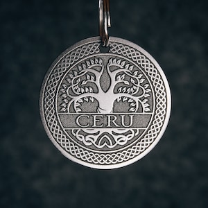 Op de afbeelding: Zilverkleurige metalen sleutelhanger met een Keltisch knoopontwerp en een afbeelding van een levensboom. Het woord "CERU" is gegraveerd op de sleutelhanger.