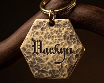 六角形の形状と濃い色の彫刻が施された、手作りの槌目加工の真鍮製ネームキーホルダー。