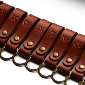 Può includere: Un set di otto portachiavi in ​​cuoio marrone con anelli in ottone. Ogni portachiavi ha un'iniziale di nome diversa impressa su di esso: SN, LC, CK, JM, JP, NF, CS.